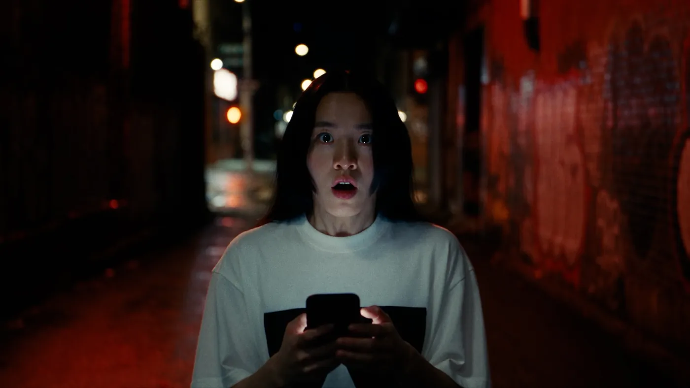 A girl in a rain-lit alley staring at her phone in shock